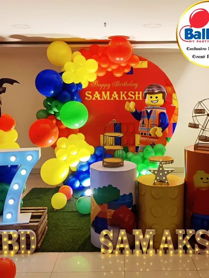 A primary-colored LEGO-themed 7th birthday party. The setup includes LEGO Batman and Emmet cutouts, LEGO-brick plinths, and a 'SAMAKSH' marquee.