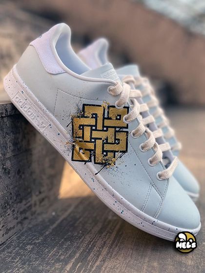 A side view of our custom Adidas Stan Smith featuring a golden, geometric Karma symbol with black paint splatters.