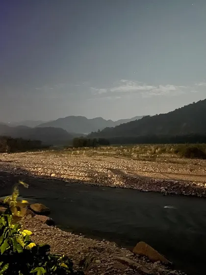 The landscape of Jim Corbett at night, illuminated by moonlight. The river and mountains are visible in the soft, ethereal light, creating a mysterious and serene scene.