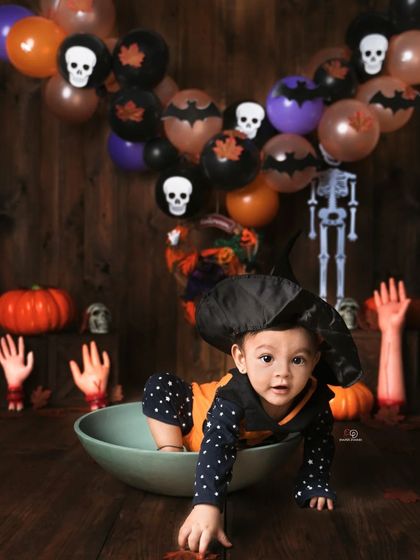 A spooky and fun Halloween-themed photoshoot. This baby, dressed in a witch hat and pumpkin outfit, crawls out of a bowl amidst pumpkins, skeletons, and other festive decorations.