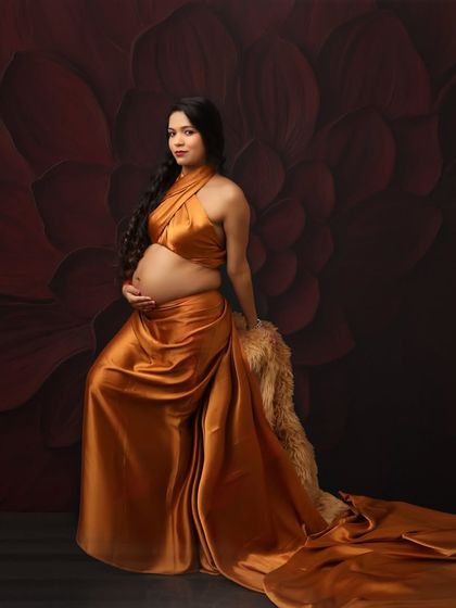 A regal portrait in a golden satin drape. The mom-to-be is seated against a rich, floral backdrop, looking like a goddess from a classical painting.