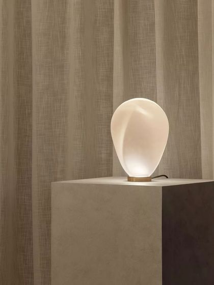 The white glass Solace table lamp emits a soft, diffused glow, its organic form beautifully illuminated against a textured curtain.