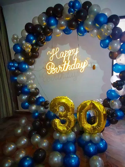 A 30th birthday celebration featuring a full balloon ring in blue, black, and silver. The setup is completed with large gold foil '30' numbers and a 'Happy Birthday' neon sign.