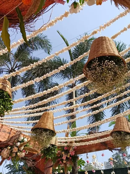 A close-up of the rustic wooden mandap, showing the detail of the hanging wicker baskets filled with baby's breath against the clear blue sky.