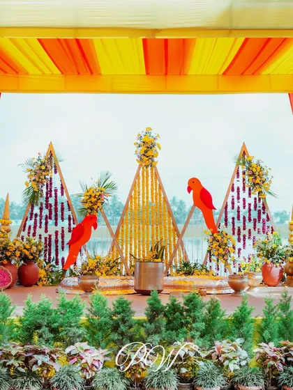 A vibrant Haldi stage with a backdrop of marigolds, geometric patterns, and parrot motifs, creating a bright and festive setting.