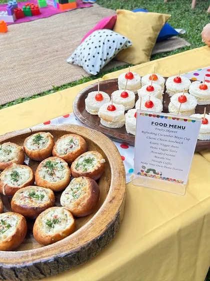 A close-up of the kid-friendly food table, showing mini veggie buns and cucumber mayo sandwiches. The printed menu adds a professional and personalized touch.