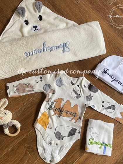 A complete and cozy set for a newborn. This collection includes a personalized animal-themed onesie, a hooded towel with bear ears, a custom cap, and a soft rattle toy for baby Shaurya.