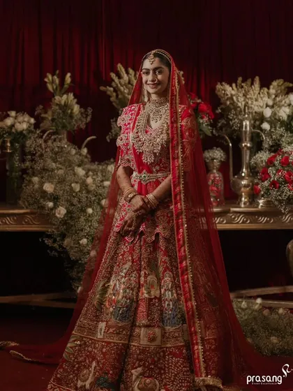 The bride stands as a portrait of elegance. The rich red drapes and abundant floral arrangements in white and red create a classic, luxurious setting that complements her traditional attire perfectly.