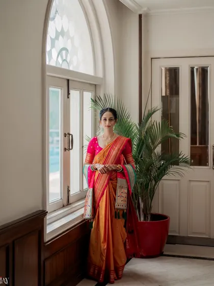 A full-length portrait of the bride in a beautiful Paithani saree, standing by a large window. The natural light from the window illuminates her, highlighting the rich colors and texture of the traditional silk saree.