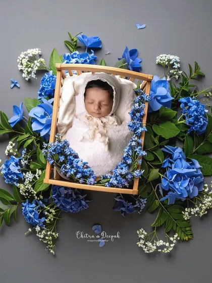 An artistic overhead shot of a sleeping newborn in a tiny wooden crib, surrounded by a bed of fresh blue and white flowers. This style of newborn photography creates a beautiful, fine-art feel.