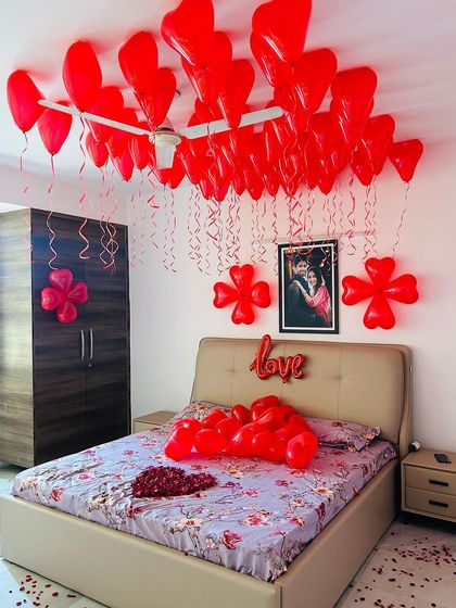A full view of a romantic room setup, combining ceiling balloons, wall decor, a "love" sign, and a heart of rose petals on the bed.