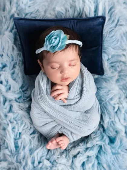 A beautifully swaddled newborn girl sleeping on a soft blue fur rug. The delicate blue floral headband and large bow add a touch of elegance to this peaceful portrait.