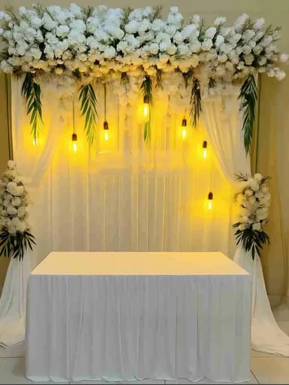 A simple and elegant backdrop for a home-based ceremony or puja, with white drapes, a floral header, and warm hanging bulbs.