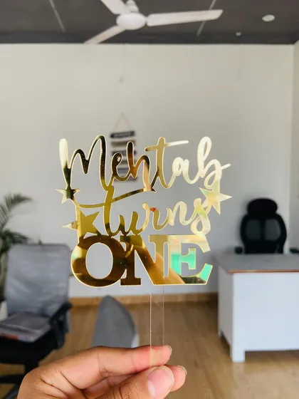 A "Mehtab turns ONE" topper held in an office setting, showing its versatility and shine in any environment.