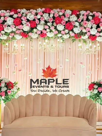 An elegant stage setup featuring a dense canopy of pink and white flowers. The soft pink drapery and classic scalloped sofa create a romantic and sophisticated setting for an engagement or reception.