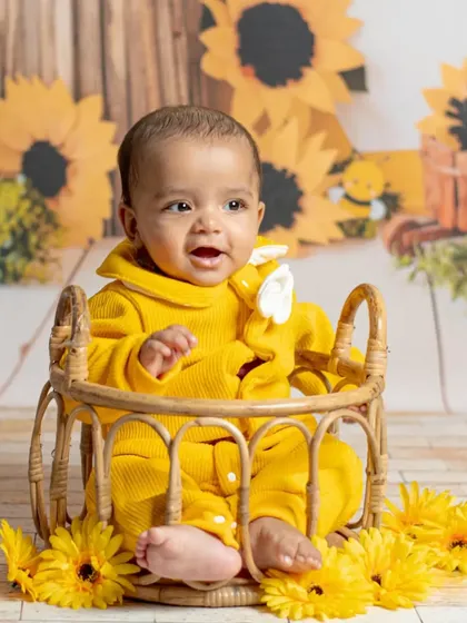 A happy baby boy in a bright yellow outfit sits in a wicker basket against a sunflower backdrop. This sitter session photo is full of joy and vibrant color.