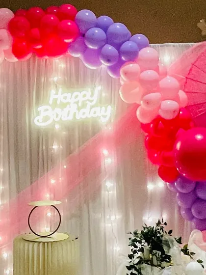 The main backdrop featuring a 'Happy Birthday' neon sign, a balloon arch in shades of pink and purple, and soft, flowing drapes with fairy lights.