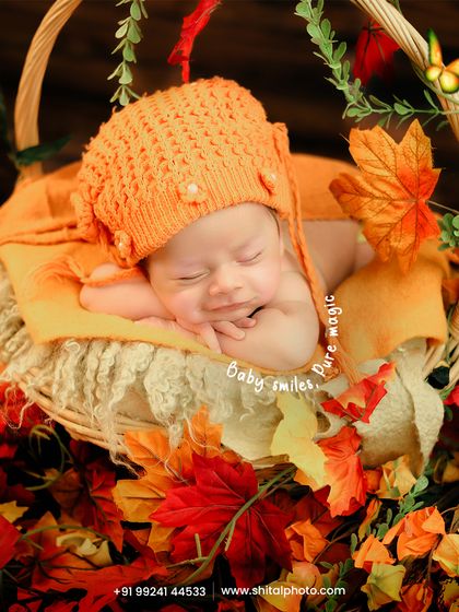 An adorable autumn-themed setup. The baby, wearing a cute orange knitted hat, smiles in their sleep while nestled in a basket filled with colorful autumn leaves.