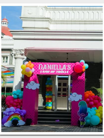 A bold and colorful entrance for "Daniella's Land of Trolls," featuring bright pink pillars, cloud cutouts, and rainbow balloon accents.