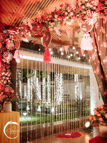 A close-up of an entrance corner, showing the layering of red floral arrangements, paper craft tassels, and sparkling bead curtains that add depth and texture to the design.