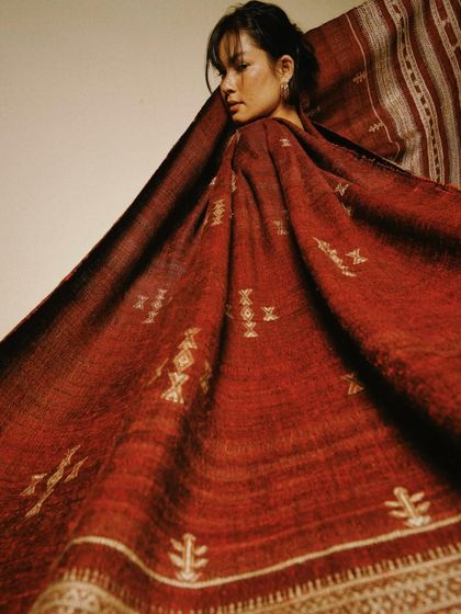 The rich, earthy red of this handwoven textile creates a dramatic and powerful image. The fabric itself becomes the main subject, enveloping the model in its beauty.