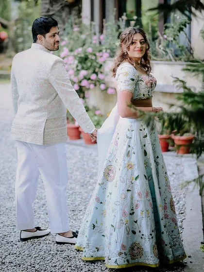 The classic "follow me to" pose from a garden engagement shoot. The bride's stunning floral lehenga and the lush greenery create a fairytale-like scene.