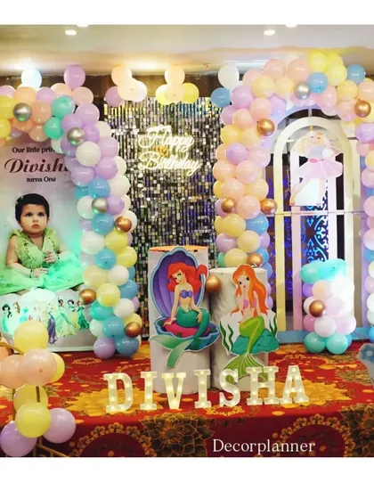 The main stage for the princess party, featuring multiple balloon arches, a shimmer backdrop, marquee letters spelling the name 'DIVISHA', and cutouts of various Disney princesses.
