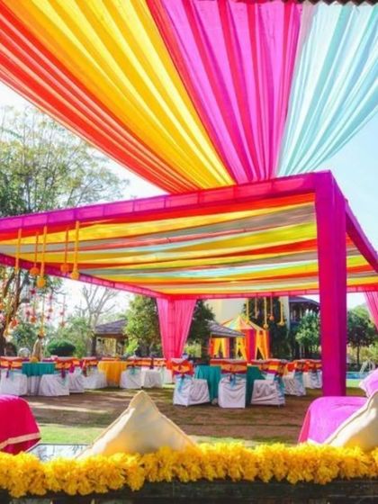 A vibrant outdoor Mehendi setup with a canopy of pink, yellow, and blue drapes. The low seating with colorful cushions is perfect for guests getting their henna done.