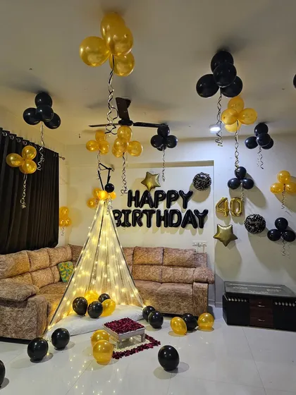 A cozy and intimate 40th birthday decoration at home. The setup includes a canopy of fairy lights, helium balloons, and a 'Happy Birthday' foil balloon banner.