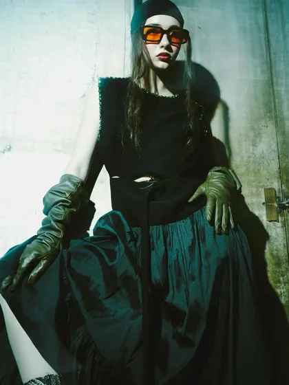The model wears bold orange sunglasses, adding a pop of color to the dark, grungy setting. The pose is confident and confrontational, fitting the avant-garde theme.