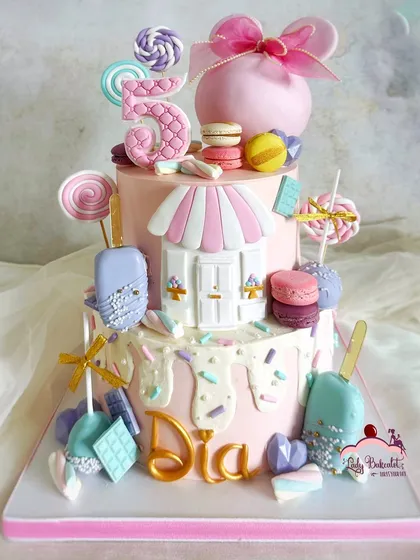 Welcome to Candyland. This cake is a dream for any sweet tooth, decorated with an assortment of edible treats like lollipops, macarons, cakesicles, and chocolate bars, all arranged to create a magical candy shop scene for a fifth birthday.