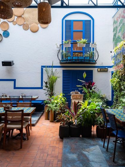 The full view of the courtyard, with its iconic blue-shuttered balcony and wall-mounted woven plates. The design is a modern interpretation of classic Greek style, creating a fresh and inviting dining area.