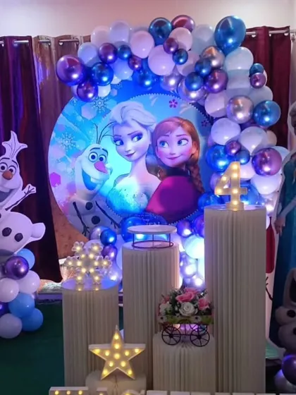 A detailed Frozen themed birthday party setup in a home. The decoration includes character standees, a circular backdrop, and a full balloon garland in shades of blue and purple.