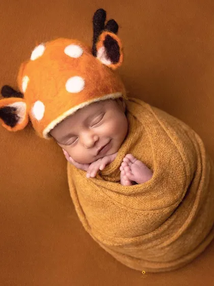 A sweet sleeping baby wearing an adorable spotted deer hat. The warm orange wrap and matching background create a cozy and whimsical woodland-themed portrait.