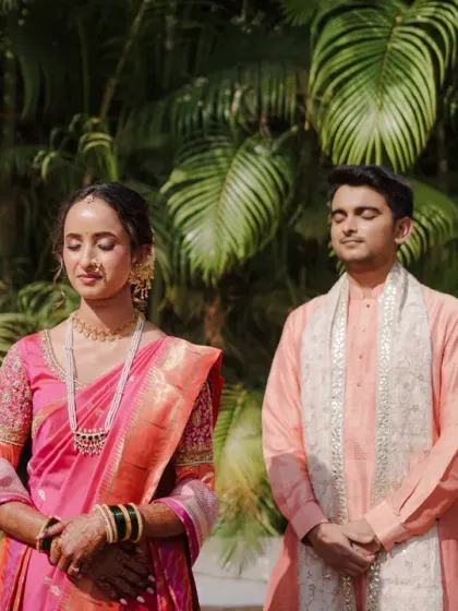 A serene moment as the Marathi bride and groom stand with their eyes closed, perhaps in prayer or quiet contemplation before a ritual. The lush green background adds to the tranquility of the scene.