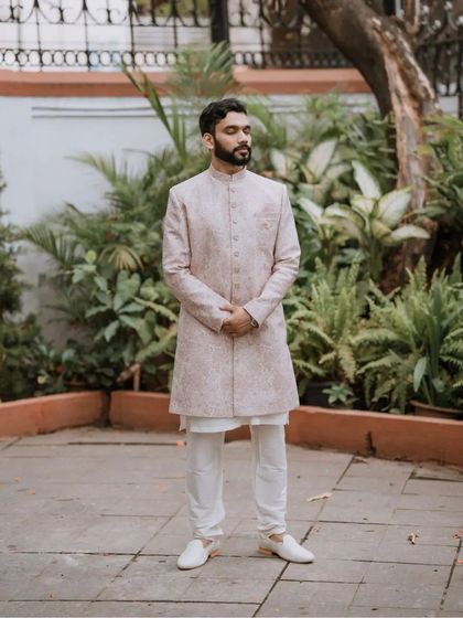 A classic portrait of the groom in a light-colored sherwani, standing in a garden setting. This image captures his calm and elegant presence on his engagement day.