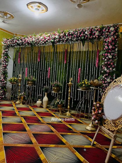 An elegant naming ceremony setup with a backdrop of hanging jasmine garlands. The stage is decorated with brass lotus stands, a traditional lamp, and a custom welcome mirror.