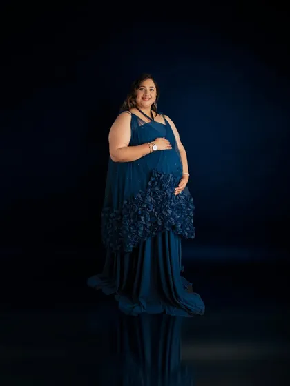A simple and elegant full-length portrait. The deep blue gown with floral appliqué stands out beautifully against the dark, reflective studio floor.