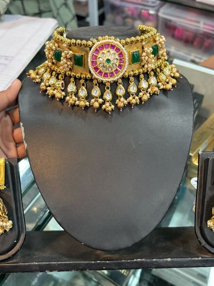 A gold-plated choker with green and pink stones, held for display. This rental set comes with matching square-shaped jhumkas.