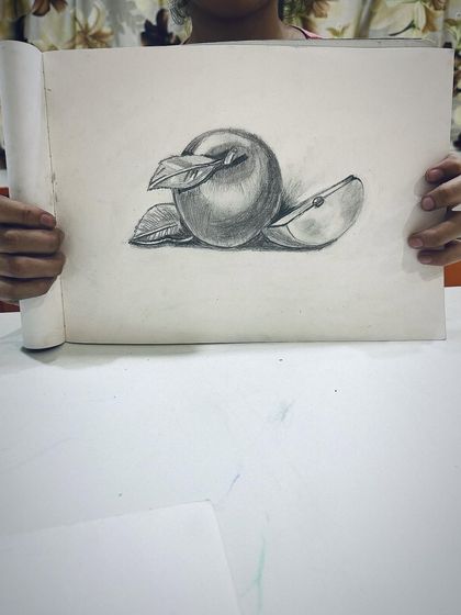 A student proudly shows their still life drawing of an apple, done with beautiful, soft shading.
