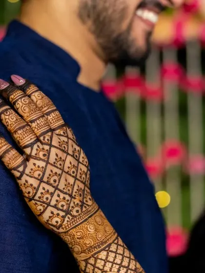 A candid moment between the couple, with the focus on the bride's hand resting on the groom's shoulder, highlighting the fresh mehendi.