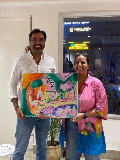 A couple holds up their vibrant and colorful paintings, one a large abstract piece and the other a smaller landscape.