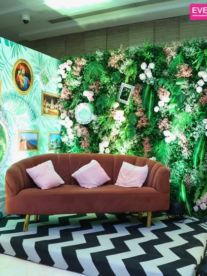 A stylish photo booth with a tropical-modern vibe. A green leaf-patterned wallpaper is paired with a real green wall, vintage frames, and a bold chevron floor, creating a multi-layered and eclectic look.