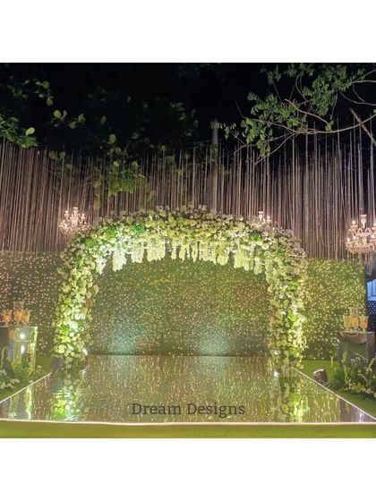 A stunning reception stage designed with a clean and modern aesthetic. The mirrored floor reflects a lush arch of white and green florals, while crystal chandeliers add a touch of timeless glamour.