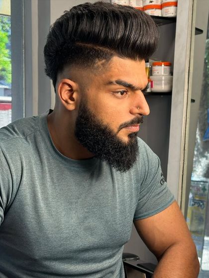 A high-volume pompadour with a skin fade and a full beard. The hair tattoo adds a unique touch.
