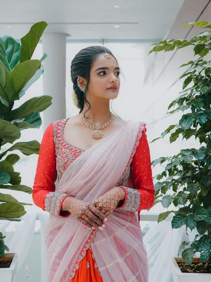 A full-length view of a custom half-saree lehenga. The bright red embroidered blouse stands out against the soft pink and orange of the skirt and dupatta.