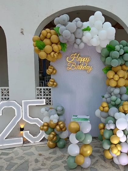 A stylish outdoor decoration for a 25th birthday. This setup includes an arched backdrop and an organic balloon garland in a sophisticated palette of sage green, gold, and white.