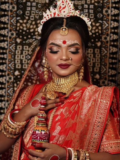 A traditional Bengali bride in a red saree, holding a 'gach kouto'. She is wearing a gold-plated choker and has the signature white and red bindi design.