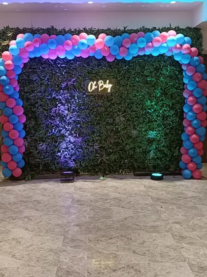 A modern "Oh Baby" entrance decor with a balloon arch against a green hedge wall.
