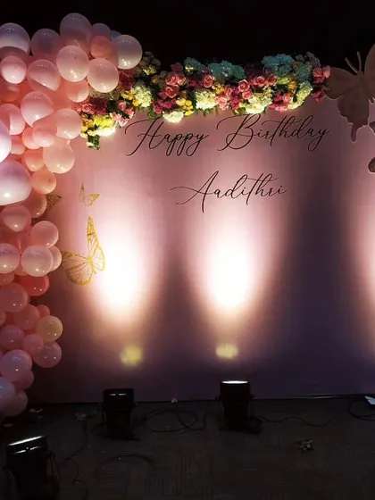 An elegant butterfly themed birthday backdrop in soft pink, accented with a pastel balloon garland and spot lighting to create a magical effect.